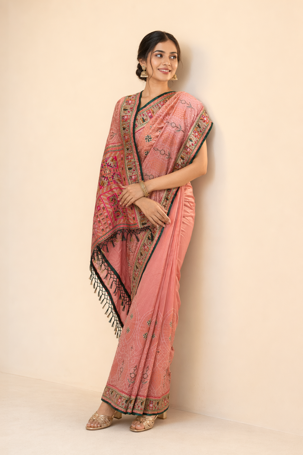 A woman modeling a pink saree with black and gold border details and a matching pink dupatta, standing against a plain beige wall.